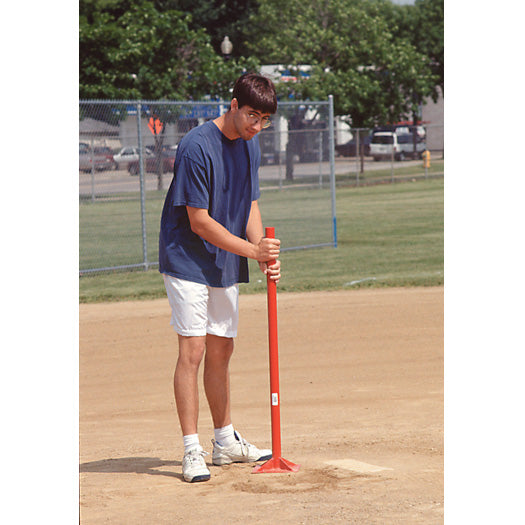A person in a blue shirt and white shorts uses the Athletic Specialties Steel Hand Tamp: TMP by Athletic Specialties to pack fill dirt on a baseball field, with grass, trees, and parked cars in the background.