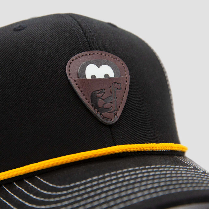 A close-up of a black cap featuring a brown leather patch shaped like a guitar pick, with a stylized B and a musical note, and a yellow cord above the brim with white stitching.