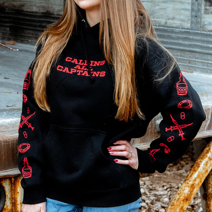 Black pullover hoodie with CALLING ALL CAPTAINS written in red font across the chest. On both sleeves there are drawings of lifesavers, beer, pills, and syringes. An individual is modeling the hoodie and standing in front of a rusted surface.