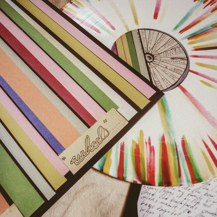 Vinyl jacket with album artwork of rainbow paper strips laying down vertically. Peeking out of the jacket is a white, yellow, red, orange, and green splattered vinyl. Both are laying on top of a surface.