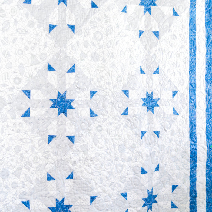 A close-up of a quilted textile featuring a white and light gray floral pattern with blue geometric star and triangle motifs, plus a blue striped border on the right edge.