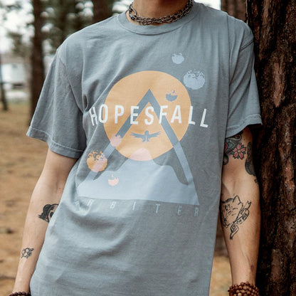 Grey short sleeve shirt with "HOPESFALL" written in white lettering across the chest. Behind that is an orange sun and a blue triangle making the shape of a mountain. There is a bird drawn over the sun in the same blue used for the mountain. Below the mountain is blue font that says "ARBITER". An individual is modeling the shirt, standing up against a tree.