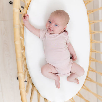 Smiling infant laying in rattan bassinet while wearing Kyte Baby Short Sleeve Bodysuits Bodysuit in Blush