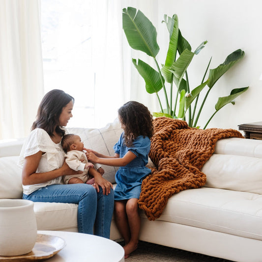 A mother and her children sit by a soft, ginger colored Saranoni chunky knit throw blanket. The large knit Saranoni blanket, is made from 100% poly microfiber and drapes beautifully. This luxury throw blanket is both trendy and durable, making it the perfect addition to any space. Ideal for home decor enthusiasts and those seeking a cuddly, soft blanket, this chunky knit blanket combines style and comfort. Keywords: chunky knit throw, soft blanket, knit blanket, large knit blanket, luxury throw