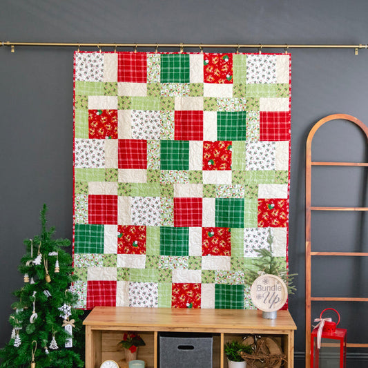 A Christmas-themed patchwork quilt hanging on a gold rod against a dark gray wall, styled with a small decorated Christmas tree, a wooden ladder, and a wooden storage bench.