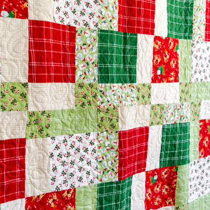 A close-up of a handmade quilt made of patchwork fabric squares in Christmas colors, featuring red, green, and white patterns including plaid, holly, pine trees, and holiday motifs.