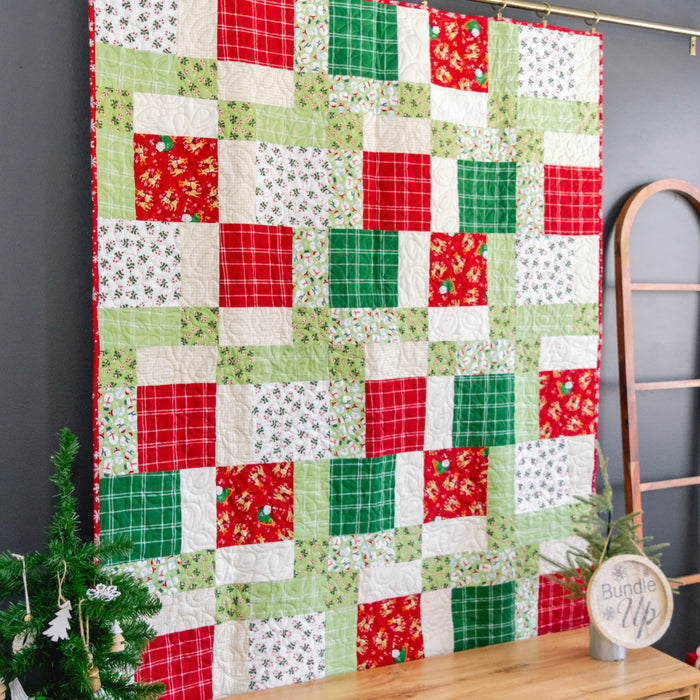 A handmade quilted patchwork blanket with a Christmas theme, featuring red, green, and white fabrics, hanging on a dark gray wall next to a wooden ladder and a small decorated tree.