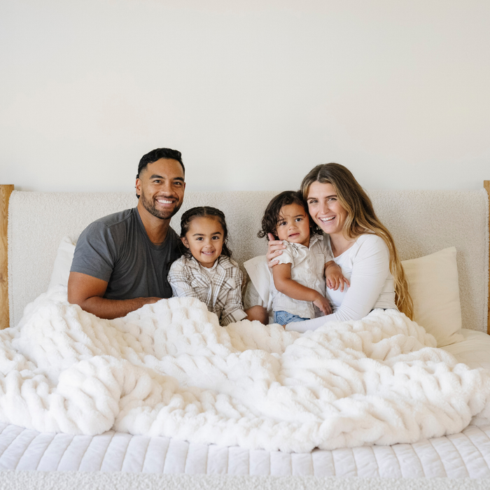 A husband and wife and their son and daughter sit under a white double ruched faux fur blanket. The soft blanket is a Saranoni blanket and a luxury blanket.