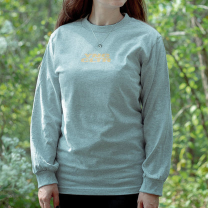 Grey long sleeve shirt with YNG CLTR in orange on the chest. On the back there is a ca drawn in orange with a triangle around it. Above that, there is text that says GODSPEED, also printed in orange. An individual is modeling the shirt, standing in front of greenery.