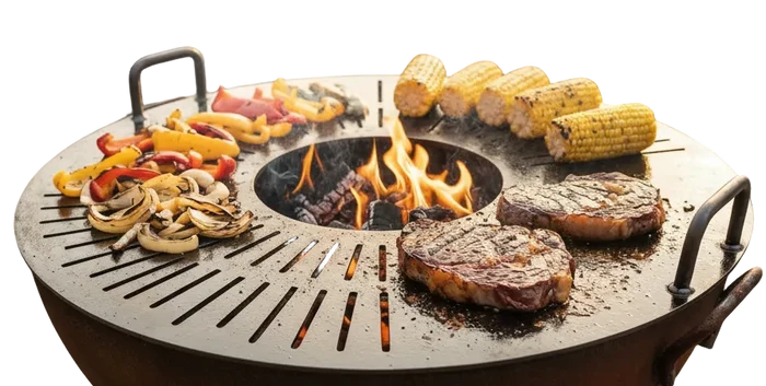 A round outdoor fire pit grill with handles sits on a gravel patio, cooking two steaks, corn on the cob, and sliced peppers and mushrooms over an open flame. Two patio chairs and stacked firewood are visible in the background of this backyard scene.