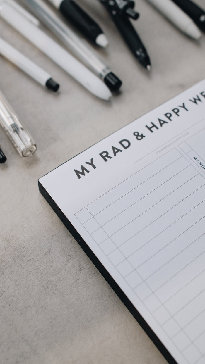 Artistic shot of the corner of a notepad with 'My Rad & Happy Writing' text and various pens on a light surface
