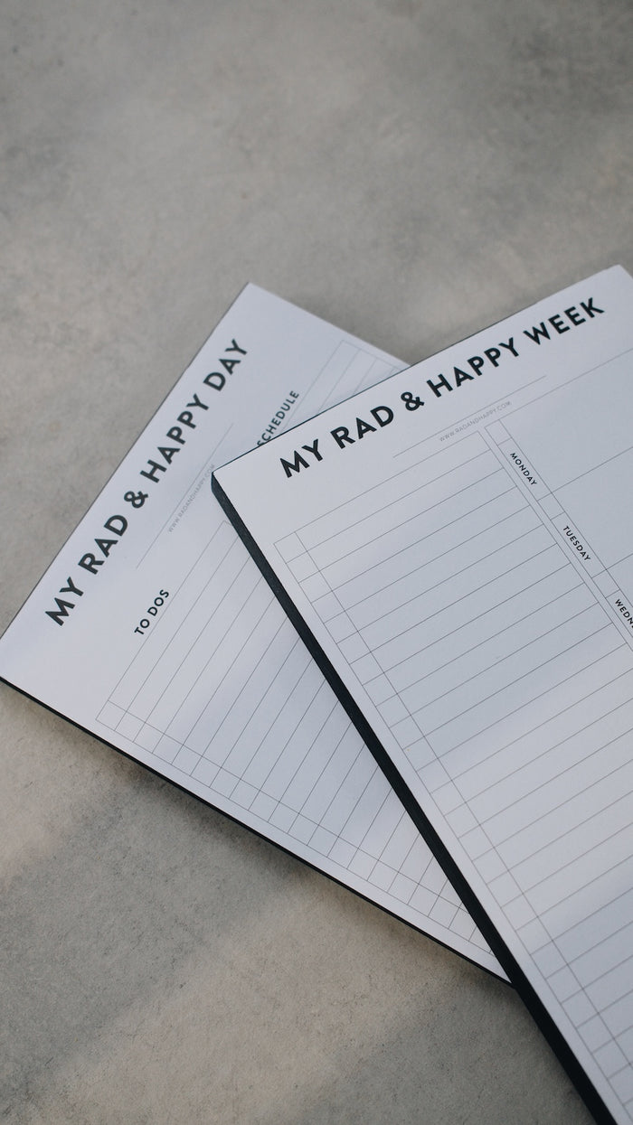 Two notepads stacked with the text "My Rad & Happy Week" on a gray surface