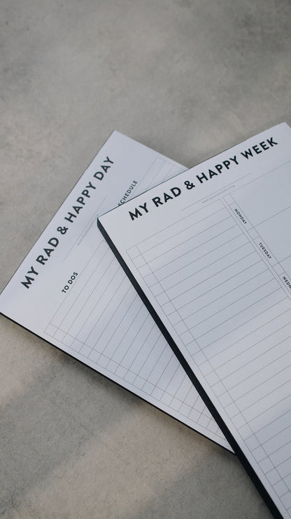 Two notepads stacked with the text "My Rad & Happy Week" on a gray surface