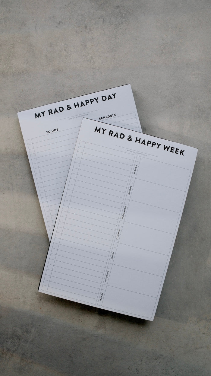 Two planner notebooks titled 'My Rad & Happy Day' and 'My Rad & Happy Week' on a gray surface.