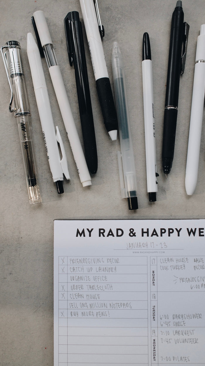Collection of pens on a concrete surface with a notepad titled 'My Rad & Happy Week'.