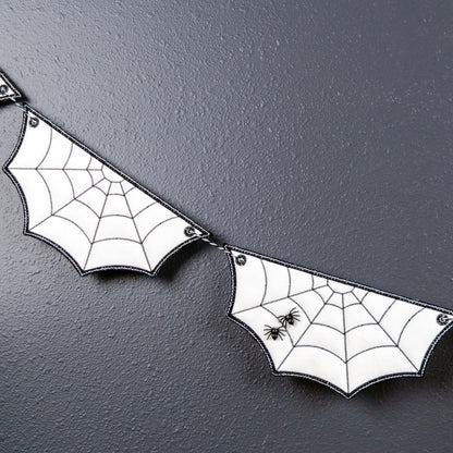 White bat-shaped banner flags with black spiderweb patterns and small black spider decorations, strung together against a dark textured wall with a small green plant branch in the corner.