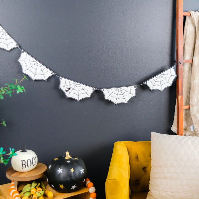 A Halloween-themed bat banner with black spiderweb designs hangs on a dark gray wall in a styled room setting with a yellow armchair, decorative pumpkins, and a plant.