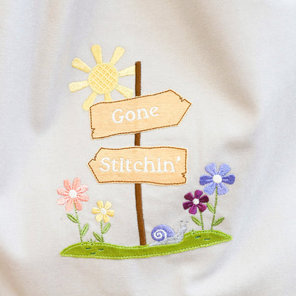 Embroidered design on a white fabric with signs and flowers, featuring the text 'Gone Stitchin'.