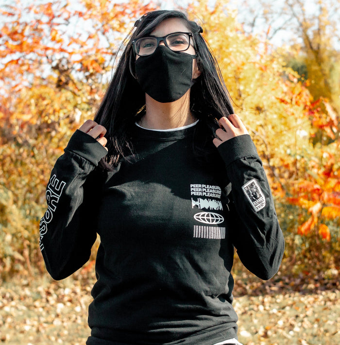 Front of black full zip hoodie. PEERPLEASURE written vertically on one sleeve in white lettering. On the top corner of the chest, there is a soundwave, globe, and barcode, and text that says PEERPLEASURE drawn in white. An individual is modeling the long sleeve standing in front of trees with leaves changing colors.
