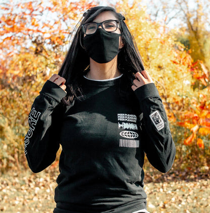 Front of black full zip hoodie. PEERPLEASURE written vertically on one sleeve in white lettering. On the top corner of the chest, there is a soundwave, globe, and barcode, and text that says PEERPLEASURE drawn in white. An individual is modeling the long sleeve standing in front of trees with leaves changing colors.
