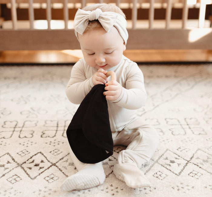 Infant playing with Kyte Baby Lovey in Midnight while wearing Zippered Footie and Headband in Oat