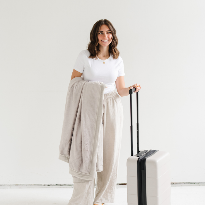 A woman holds a Feather (light gray) Lush XL Saranoni blanket, designed for oversized comfort and featuring a buttery soft, plush texture and has her other hand on a piece of luggage. This luxurious Saranoni blanket is perfect for cozy evenings, reading, or home decor. The Saranoni blanket adds warmth, style, and unmatched softness, making it an ideal gift or essential for ultimate relaxation.