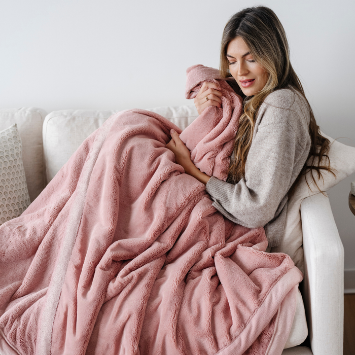 A woman sits on a couch while holding a pink blanket that is a lush blanket. The soft  blanket is a luxury blanket and a Saranoni blanket. This extra large blanket is great for relaxing, as a comfort gift, or as a home decor staple. Enjoy this cozy blanket for years to come.