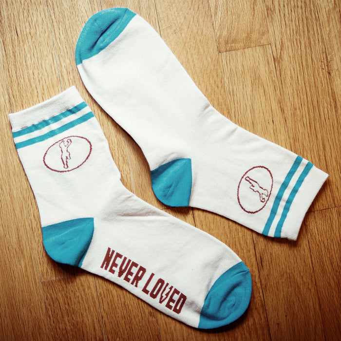Cream colored socks with blue accents and a red circular design on the top. NEVER LOVED is written in red on the bottom of the socks. Socks are laying on a wooden surface.