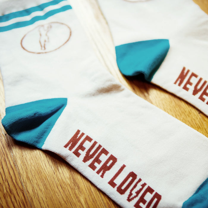 Cream colored socks with blue accents and a red circular design on the top. NEVER LOVED is written in red on the bottom of the socks. Socks are laying on a wooden surface.