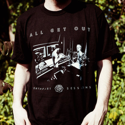 A person wears an All Get Out black Tultex T-shirt with a monochrome image of two people in a music studio, ALL GET OUT at the top, and NORTHPORT SESSIONS at the bottom. Blurred green foliage is visible in the background.