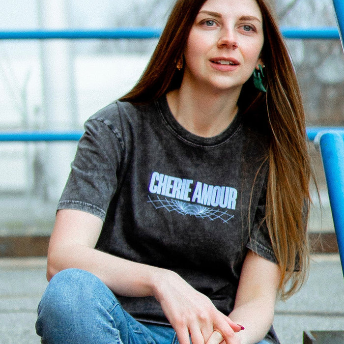 Black short sleeve shirt with CHERIE AMOUR written across the chest in periwinkle font. Underneath that is a design made of a series of curved lines. The shirt is being modeled by an individual sitting on steps with blue railings surrounding it.