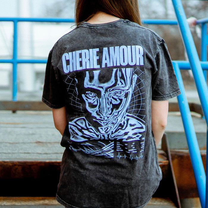 Back of black short sleeve shirt with CHERIE AMOUR written across the top in periwinkle font. Below that is a drawing of a face and the top of their shoulders. An individual is modeling the shirt and standing on stairs with blue railings surrounding them.