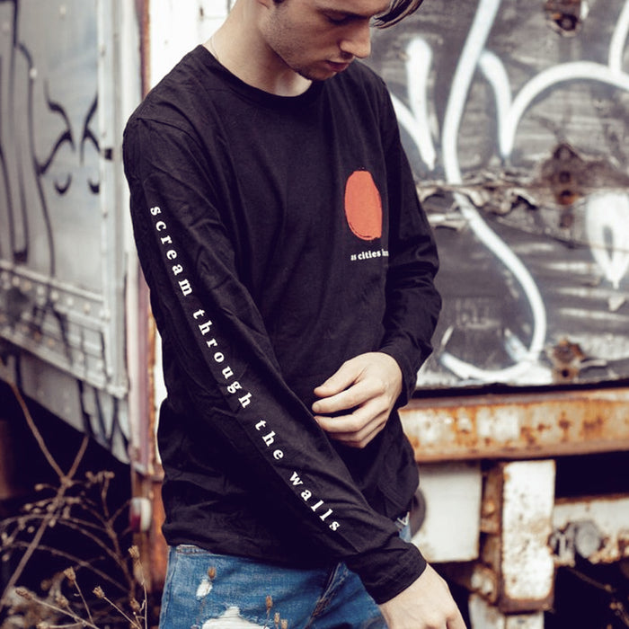 Black long sleeve shirt with a red paint dab design in the top corner. Below that is small white text that says AS CITIES BURN. Down one sleeve there is text that says SCREAM THROUGH THE WALLS. An individual is modeling the shirt and standing outside in front of a graffitied truck.