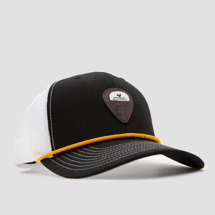 A black and white mesh trucker hat with a yellow cord above the brim and a brown leather patch featuring an animal logo on the front. The background is plain light gray.