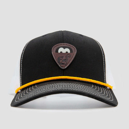 A black and white trucker hat with a yellow cord above the brim, featuring a brown patch shaped like a guitar pick with a cartoon face and guitar graphic on the front.