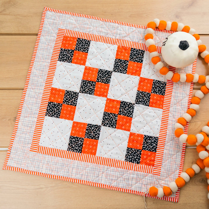 A handmade quilted baby blanket with a checkered pattern of orange, black, and white squares, styled on a light wood background next to a white pom-pom toy.