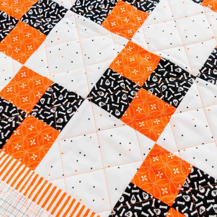 A close-up of a quilted fabric with a patchwork design featuring orange and black patterned squares on a white background, finished with an orange and white striped border.