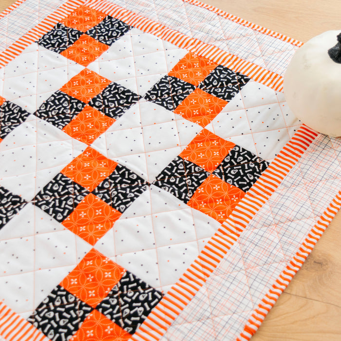 A handmade quilted table runner or placemat featuring a geometric patchwork design of orange, black, and white fabrics, styled on a light wood surface next to a small white decorative pumpkin.