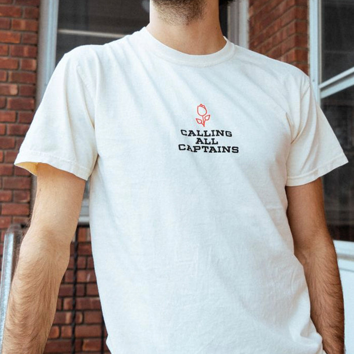 Cream colored short sleeve shirt with CALLING ALL CAPTAINS written across the chest in black font. Above the text there is a small red rose. An individual is modeling the shirt and standing in front of a brick building with two windows and a railing in the background.