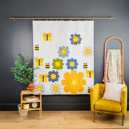 A white fabric wall hanging or curtain featuring a colorful floral and bee pattern, displayed on a gold rod in a styled room with a yellow armchair, side table, and dark gray wall.