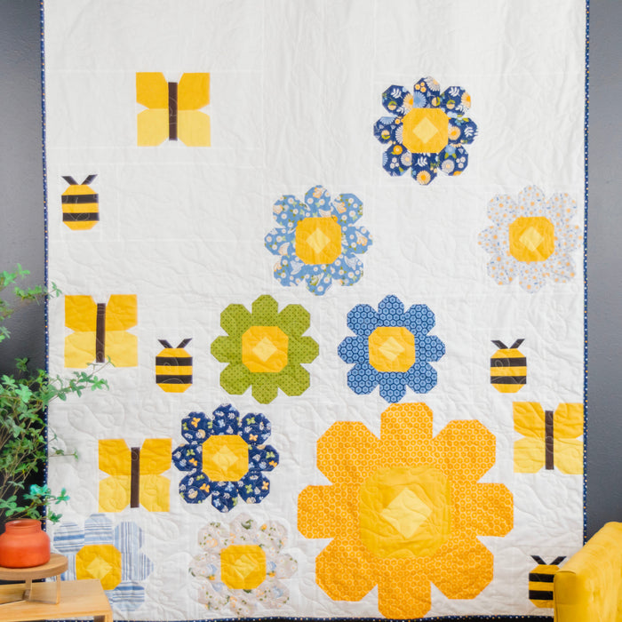 A quilted wall hanging or blanket featuring a whimsical bee and flower pattern in yellow, blue, and green on a white background, styled in a bedroom setting next to a yellow chair and a plant.