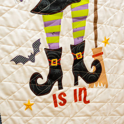A close-up of a quilted fabric wall hanging or panel with a Halloween design. The cream-colored quilt features applique legs in black and purple striped stockings, black witch boots with gold buckles, a broom, stars, and the words 'is in' in red.