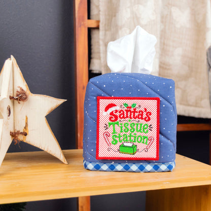 A quilted blue tissue box cover with a white tissue protruding, decorated with a red and green 'Santa's Tissue Station' patch. It sits on a wooden table next to a wooden star ornament, with a Christmas tree and a chair in the background.
