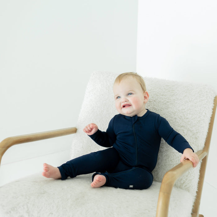 Smiling baby sitting on a white chair while wearing a Zippered Romper in Navy